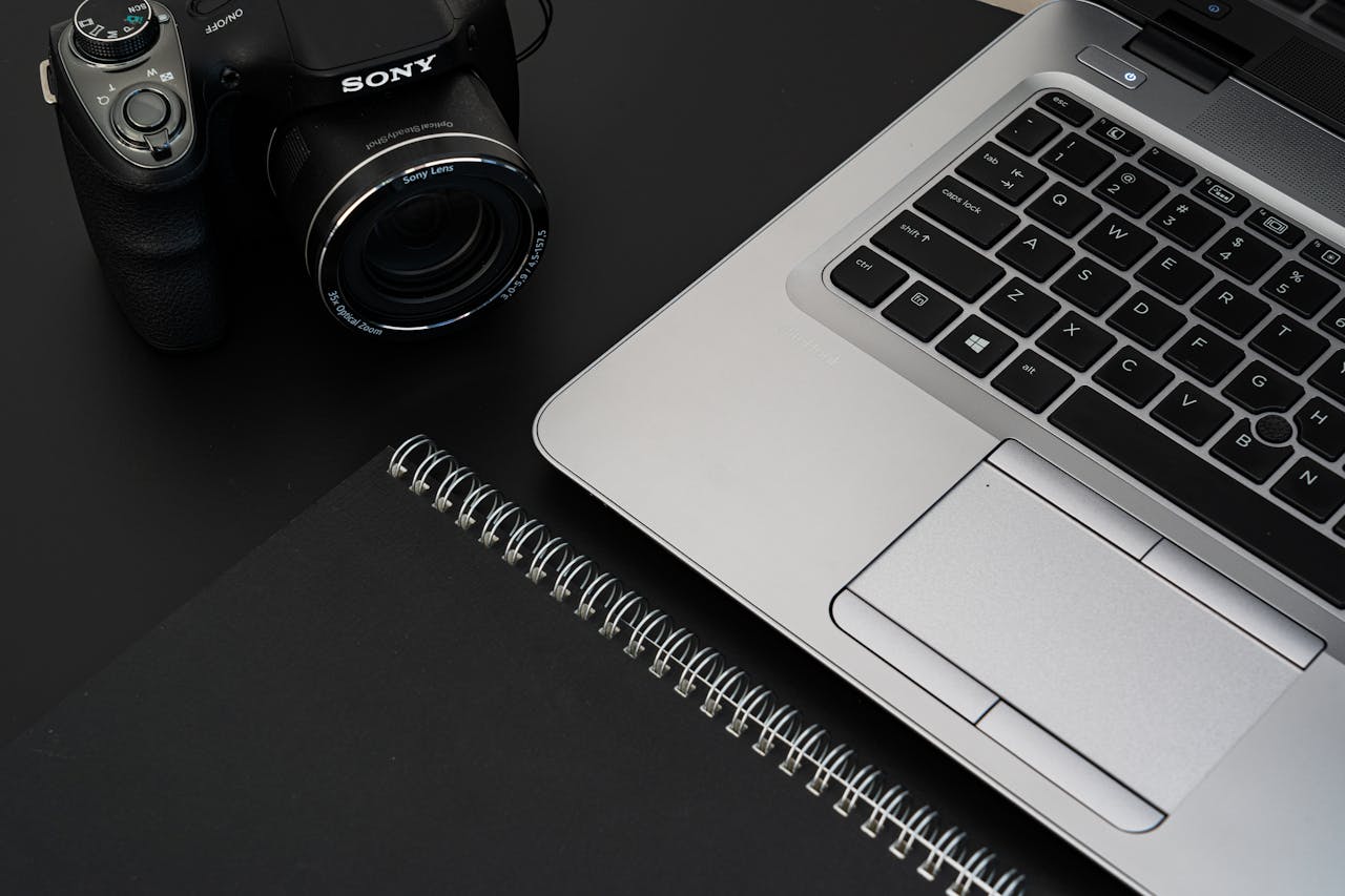 A stylish office desk featuring a laptop, camera, and spiral notebook on a dark surface.
